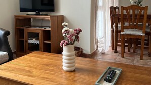 Una sala de estar con una mesa de centro de madera, un televisor sobre un mueble, un jarrón con flores y una zona de comedor con sillas y una mesa.