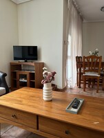 Una sala de estar con una mesa de centro de madera, un televisor sobre un mueble, un jarrón con flores y una zona de comedor con sillas y una mesa.