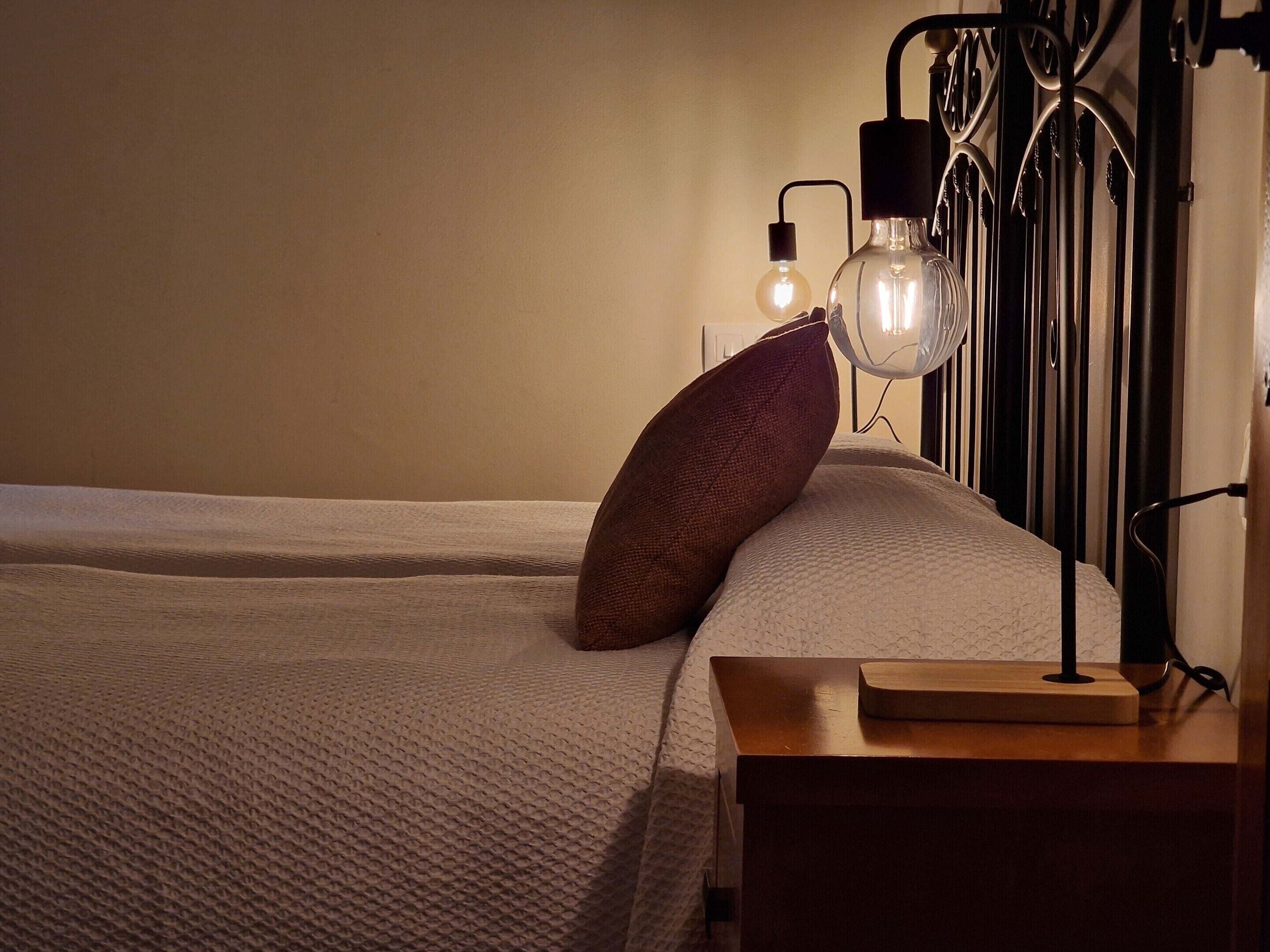 A neatly made bed with a textured white duvet, a single brown pillow, and a bedside table with a book and a lamp.