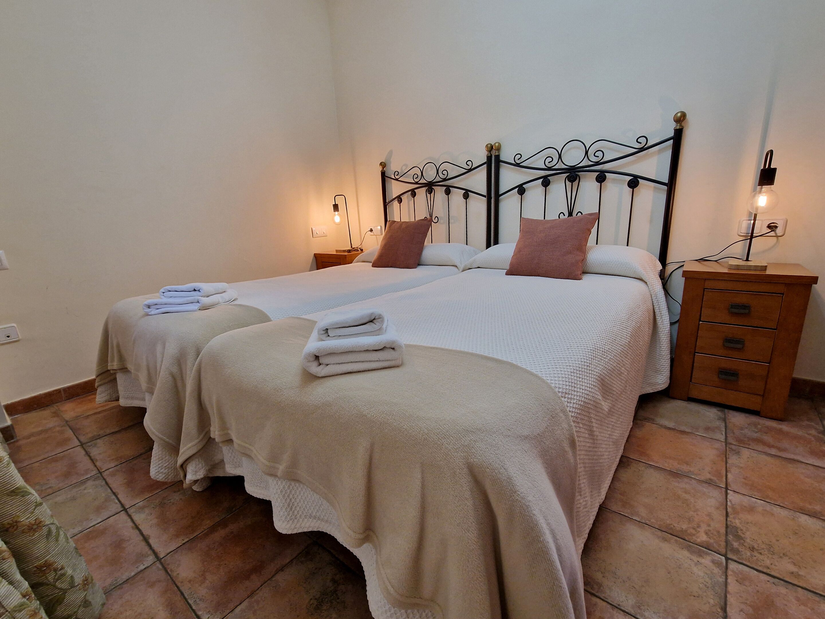 A hotel room with two beds, each with a white bedspread and pillows, a wooden nightstand with a lamp, and a tiled floor.