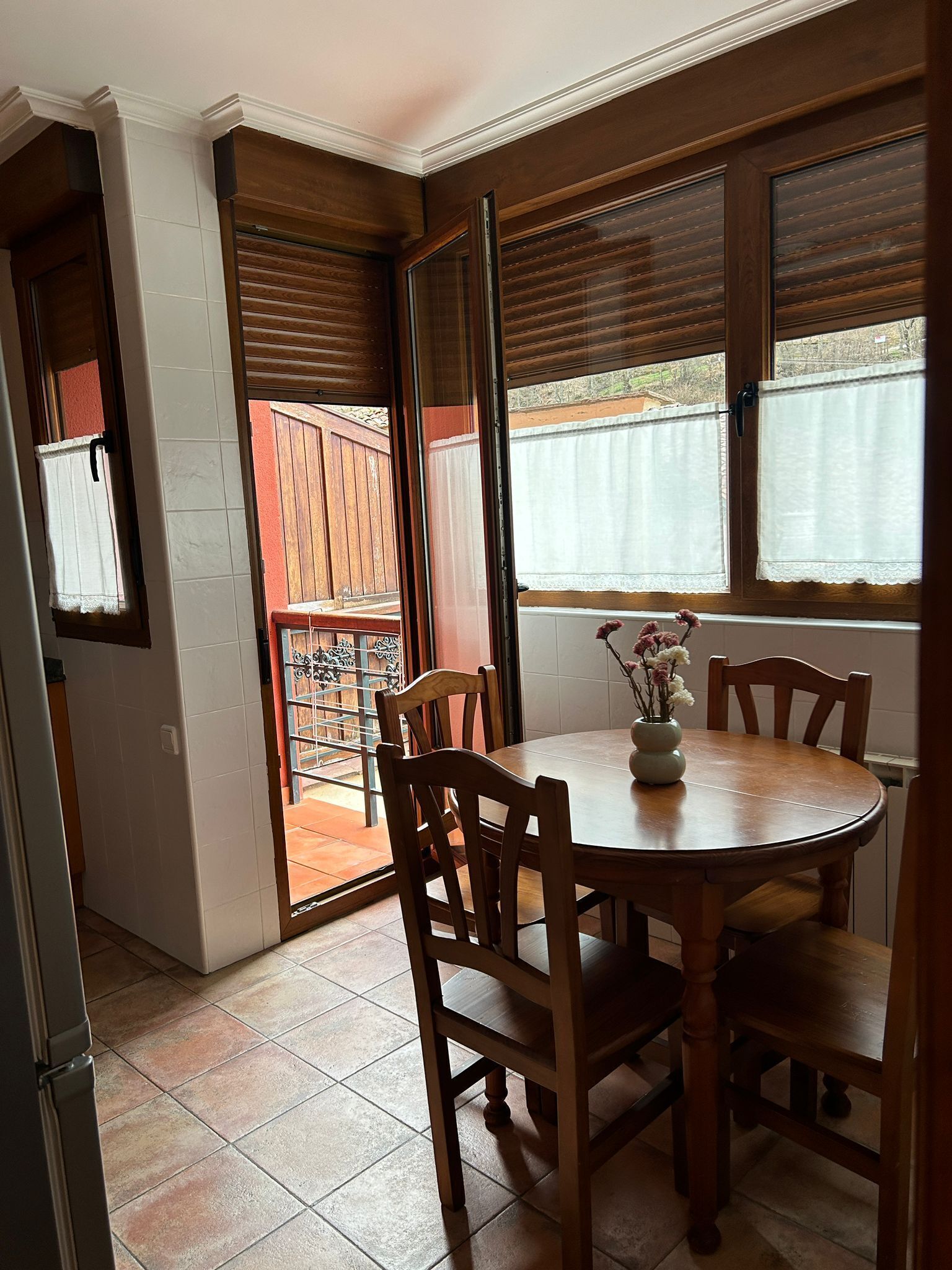 A dining area with a round wooden table, four chairs, and a vase with flowers. There are two windows with blinds, a door leading to a balcony, and a tiled floor.