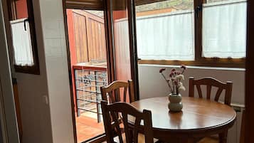 A dining area with a round wooden table, four chairs, and a vase with flowers. There are two windows with blinds, a door leading to a balcony, and a tiled floor.