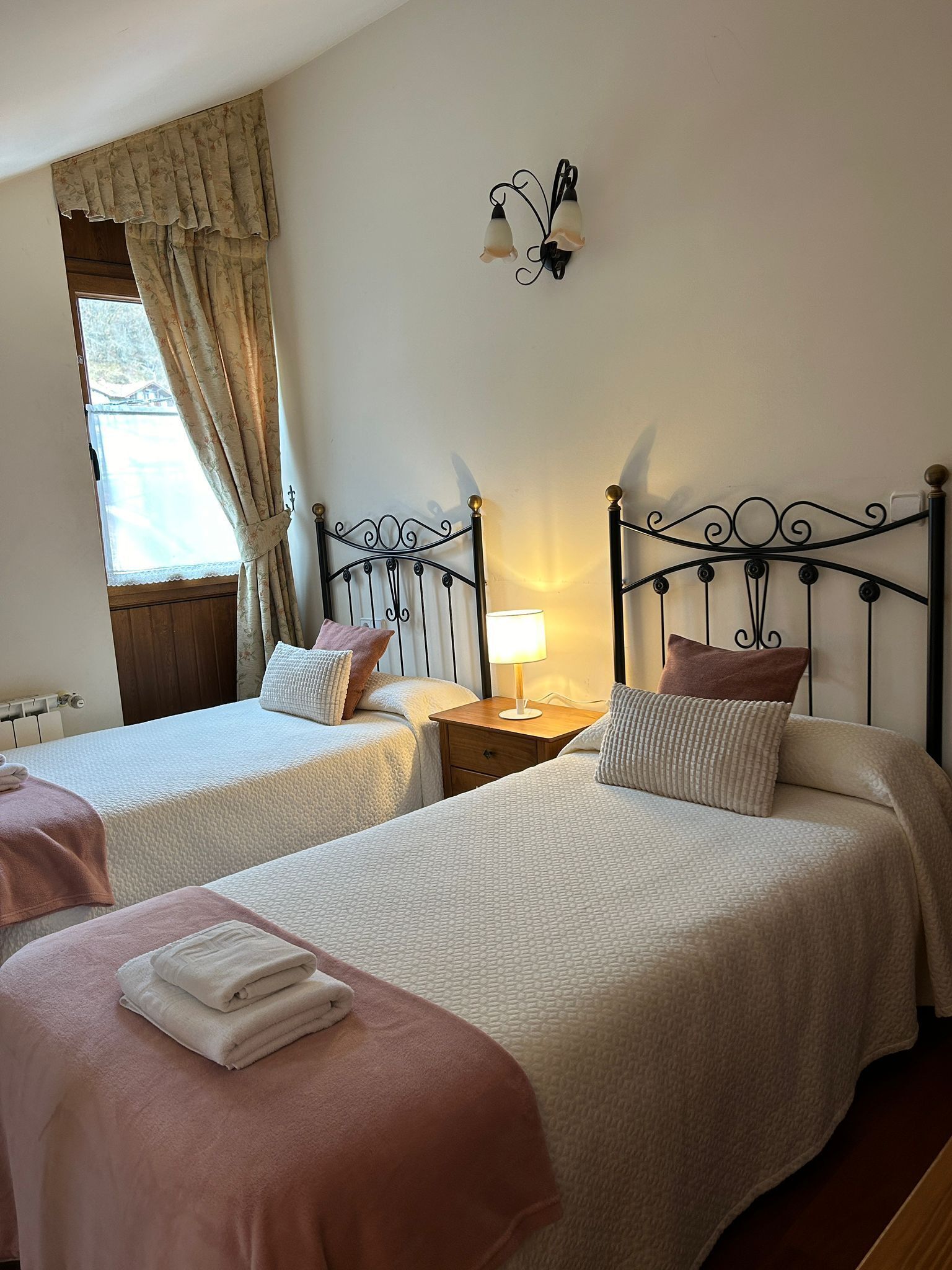 A room with two beds, each with a pillow and a folded towel. There is a nightstand with a lamp between the beds. A window with curtains is visible, and a wall-mounted light fixture is above the beds.