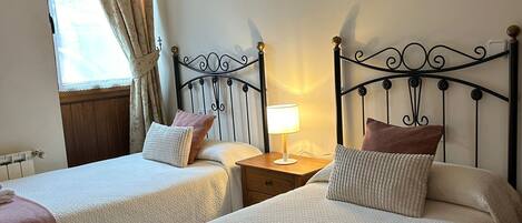 A room with two beds, each with a pillow and a folded towel. There is a nightstand with a lamp between the beds. A window with curtains is visible, and a wall-mounted light fixture is above the beds.