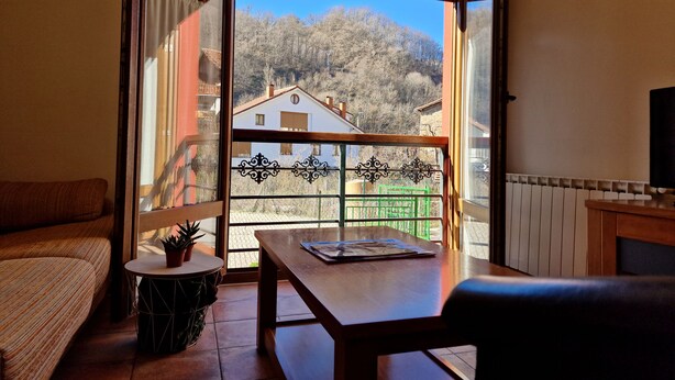 Un balcón con vistas a casas y árboles, una mesa de madera, una silla y una mesita redonda con una planta.