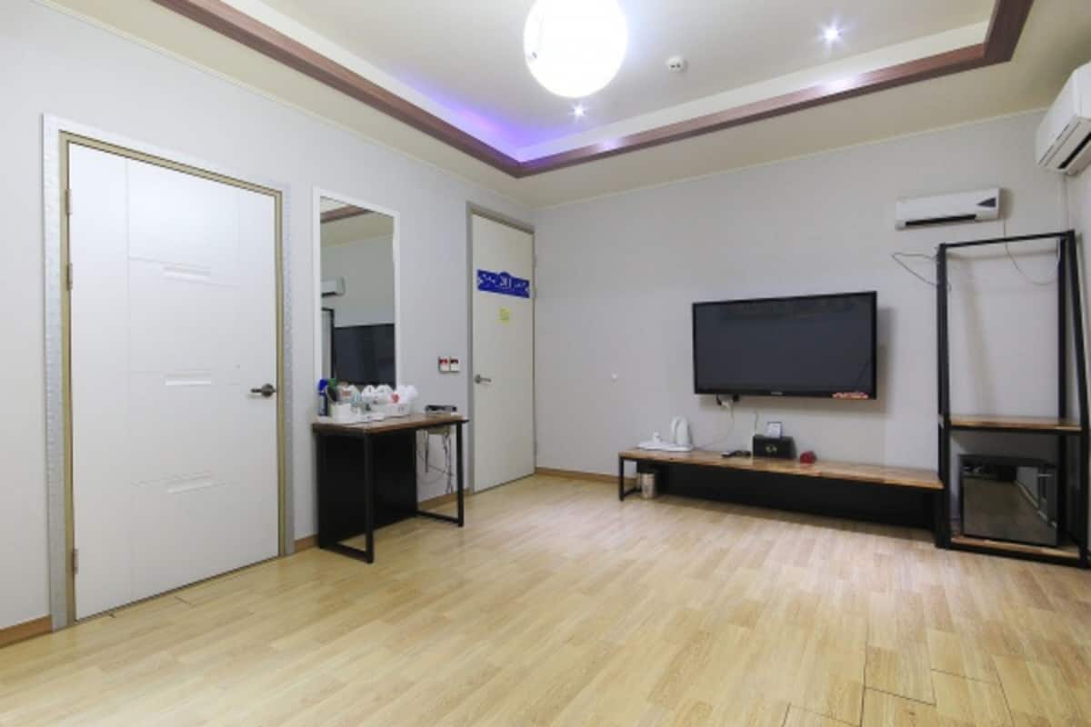 A room with a wooden floor, white walls, a flat-screen TV mounted on the wall, a small table with a chair, and a door.
