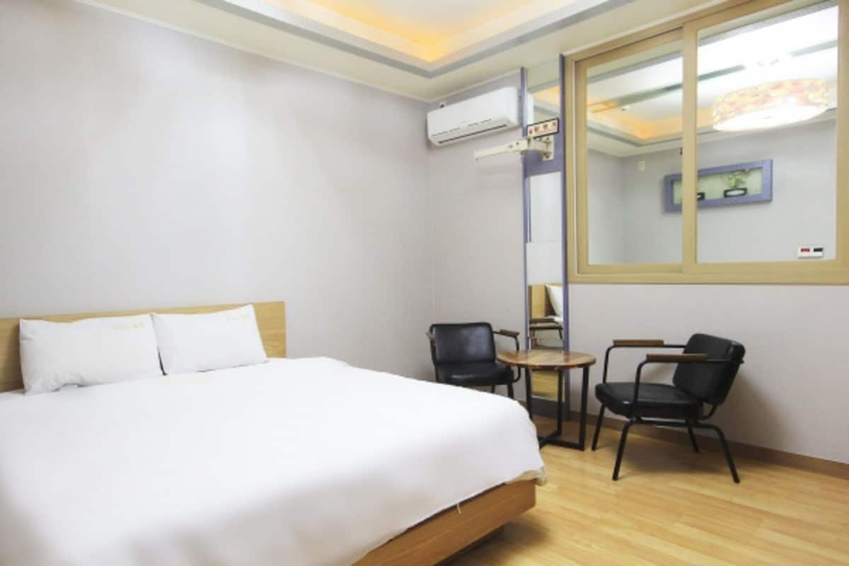 A hotel room with a large bed, two chairs, a small table, and an air conditioning unit.