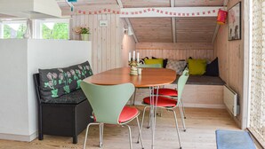 Un acogedor interior de cabaña con techo de madera, una mesa redonda de madera, sillas verdes y rojas, un sofá negro con cojines estampados y una ventana pequeña con vistas a la vegetación.