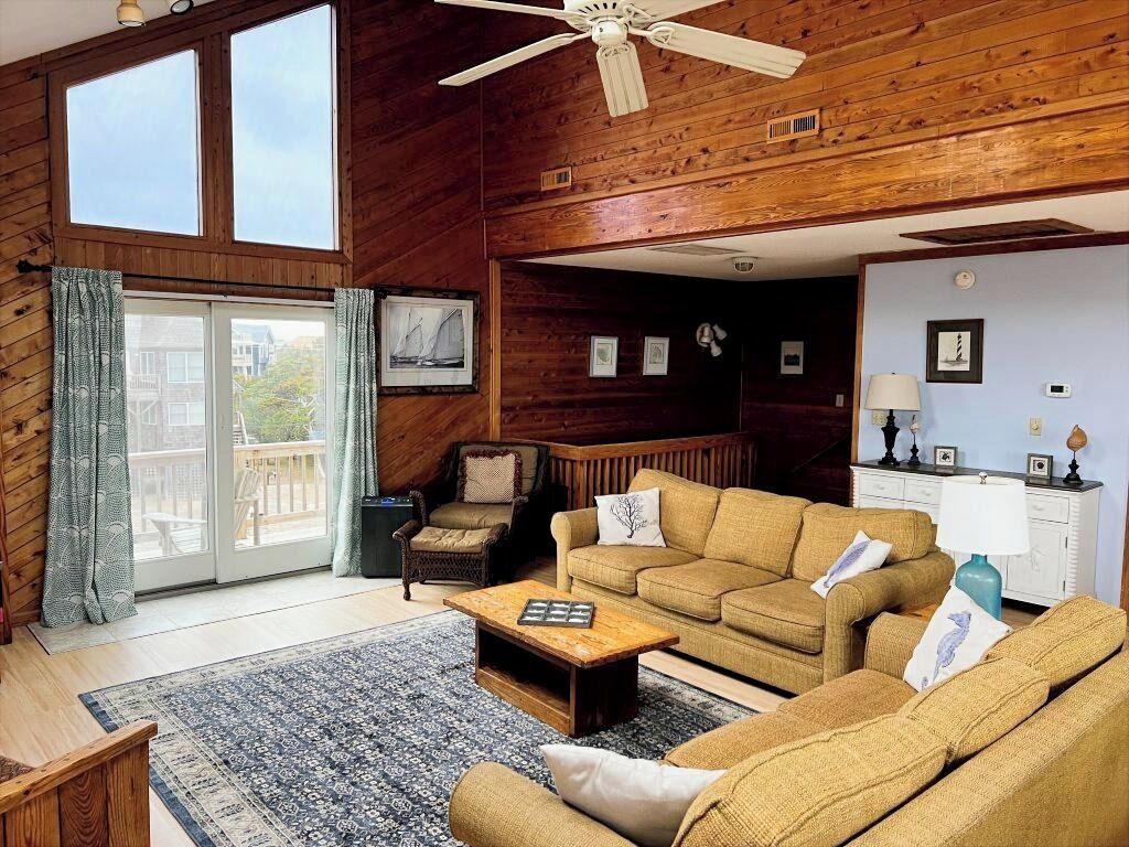 A cozy living room with wooden walls, a ceiling fan, a large window, and a sliding glass door leading to an outdoor area.