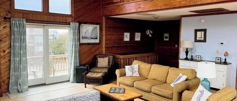 A cozy living room with wooden walls, a ceiling fan, a large window, and a sliding glass door leading to an outdoor area.