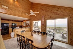 Enjoy meals at the large dining table surrounded by comfortable chairs, with a balcony offering a view of the beach.