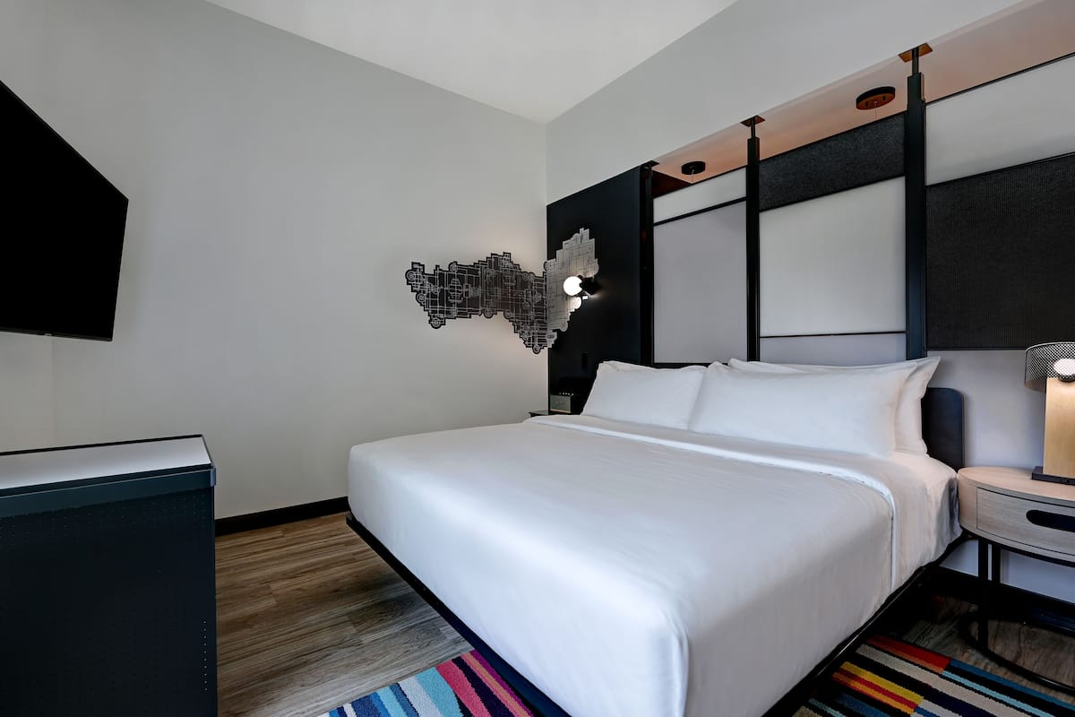 A modern hotel room with a large bed, a flat-screen TV mounted on the wall, a desk, and a unique wall art piece.