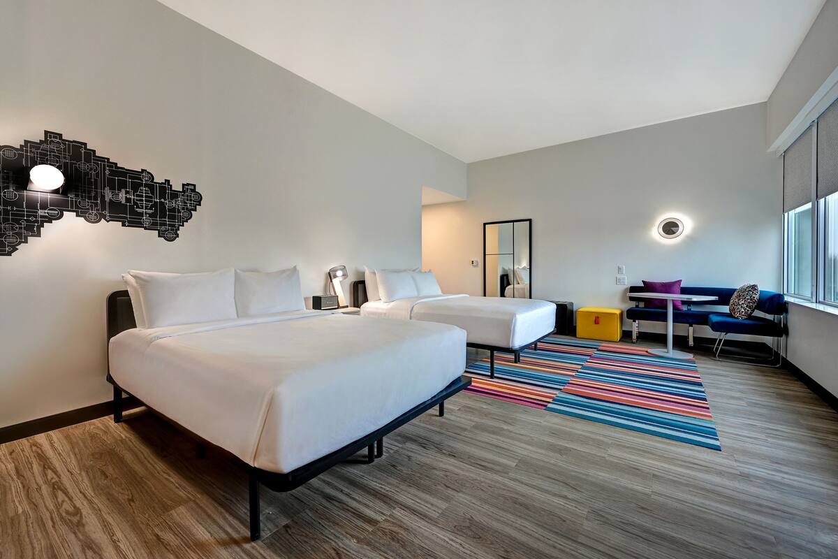A modern hotel room with two beds, a desk, a chair, a colorful striped rug, and a unique wall light fixture.