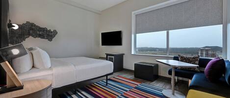 Quarto de hotel moderno com cama, escrivaninha, sofá, televisão e uma grande janela com vista para a cidade.