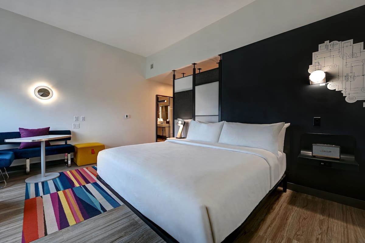 A modern hotel room with a large bed, a desk, a colorful striped rug, and a unique wall art piece.