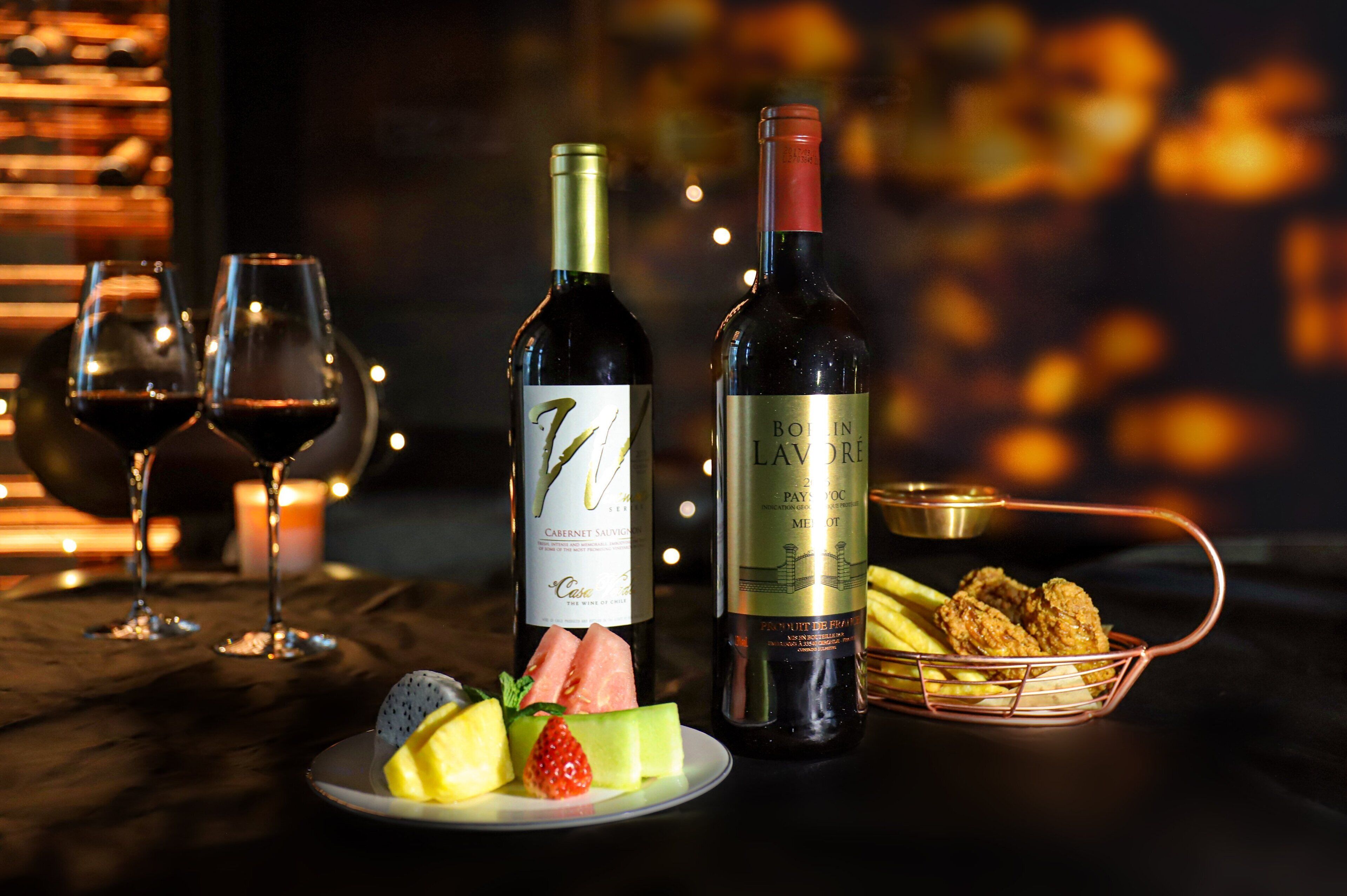 Deux bouteilles de vin, une assiette de fruits et un panier de victuailles sont posés sur une table, avec des verres à vin et des bougies en arrière-plan.
