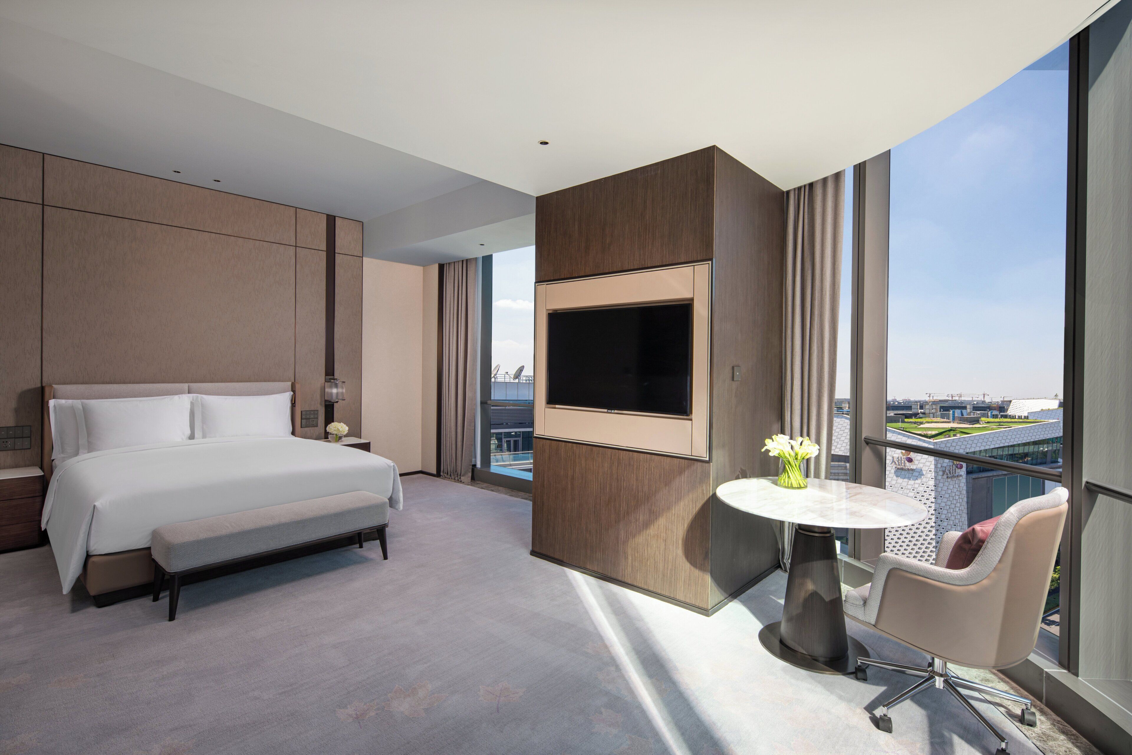 Een moderne hotelkamer met een groot bed, een flatscreen televisie aan de muur, een klein rond tafeltje met een bloemenvaas en een stoel.