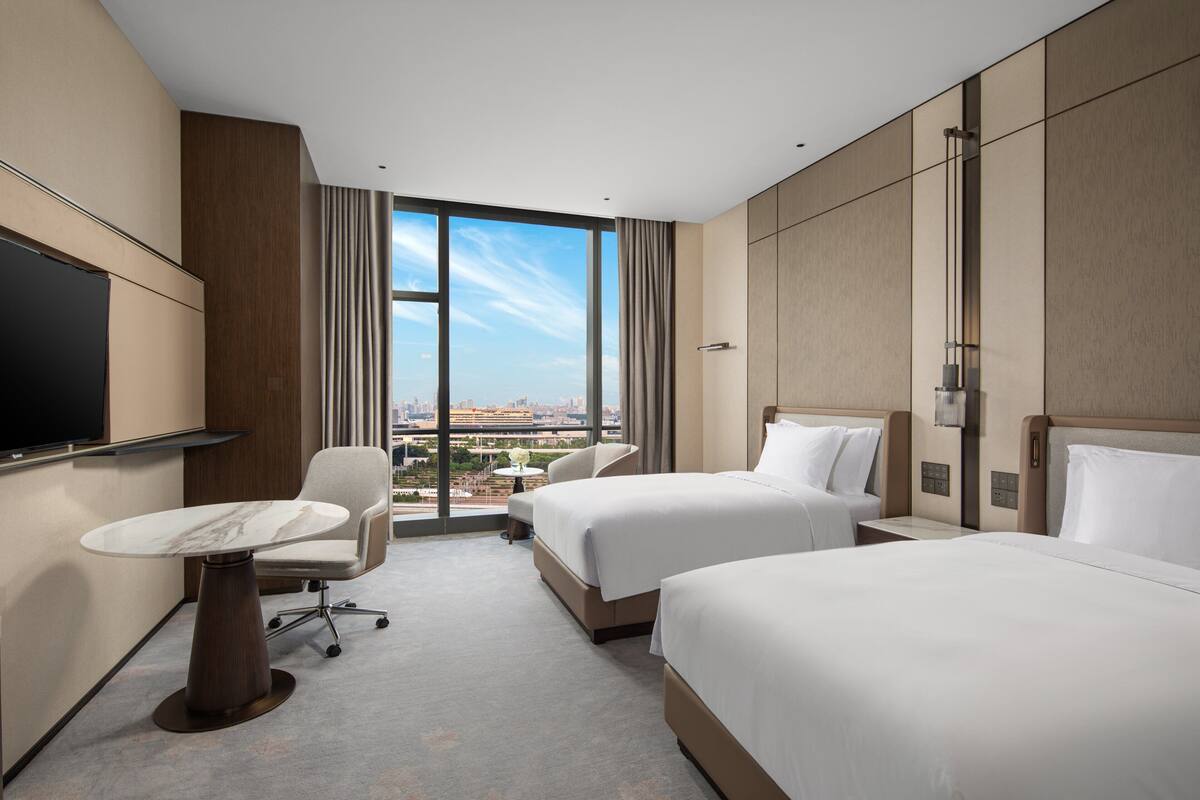 Ein Hotelzimmer mit zwei Betten, einem Schreibtisch, einem Stuhl und einem großen Fenster mit Blick auf die Stadt.