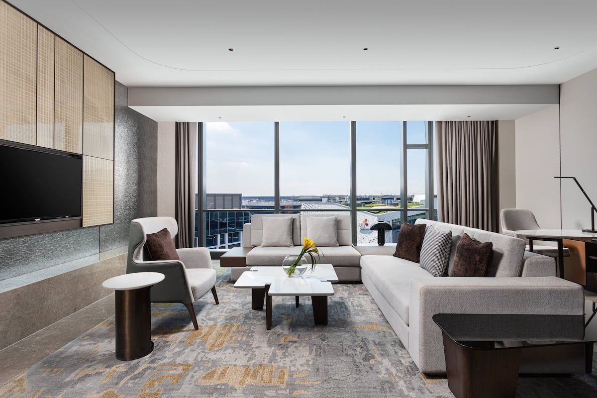Ein modernes Hotelzimmer mit einer großen Couch, Sesseln, einem Couchtisch und einem Flachbildfernseher. Große Fenster bieten Blick auf die Stadt.