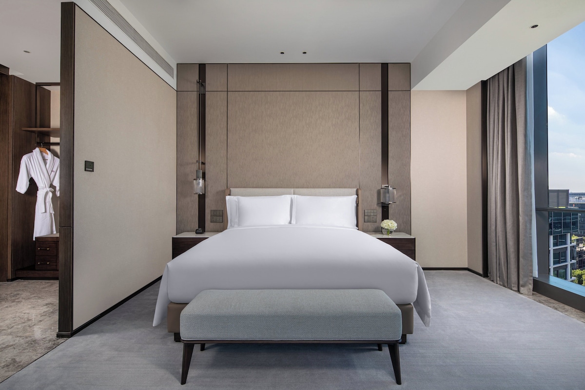 Una habitación de hotel moderna con una cama grande, mesitas de noche, un perchero para bata y vistas a la ciudad.