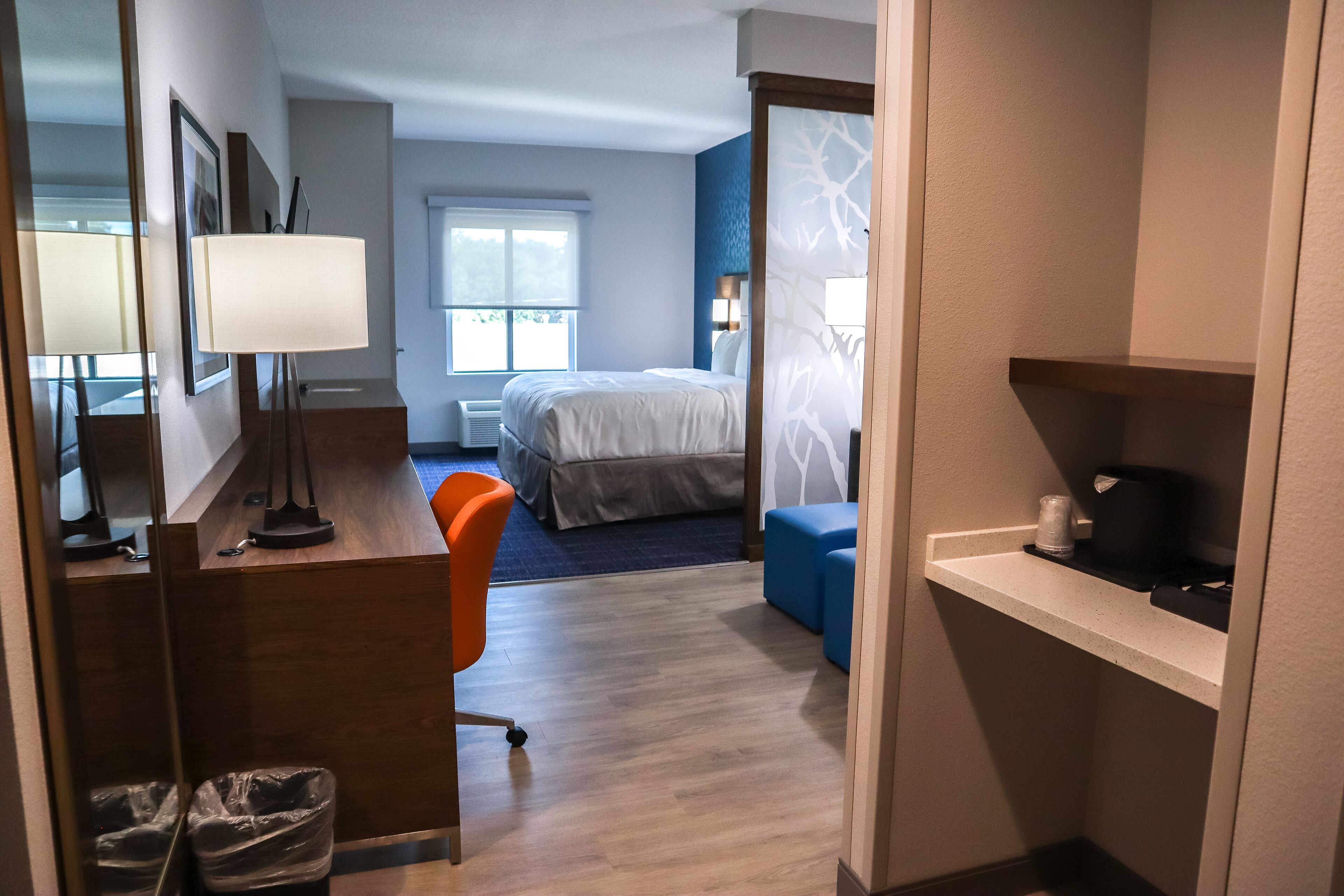 A hotel room with a bed, a desk with a lamp, a chair, a small table, and a shelf with a coffee maker.