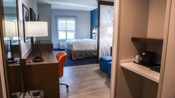 A hotel room with a bed, a desk with a lamp, a chair, a small table, and a shelf with a coffee maker.