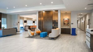 A modern hotel lobby with a reception desk, seating area with chairs and ottomans, a large artwork, and a corridor leading to other rooms.