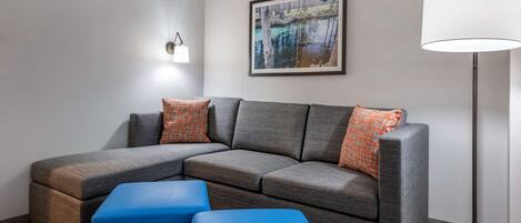Una sala moderna con un sofá modular, dos otomanas azules, una lámpara de pie y un cuadro enmarcado en la pared.