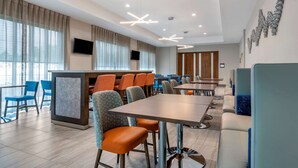 A modern hotel lobby with a long dining table, multiple seating areas, and a large window with curtains.