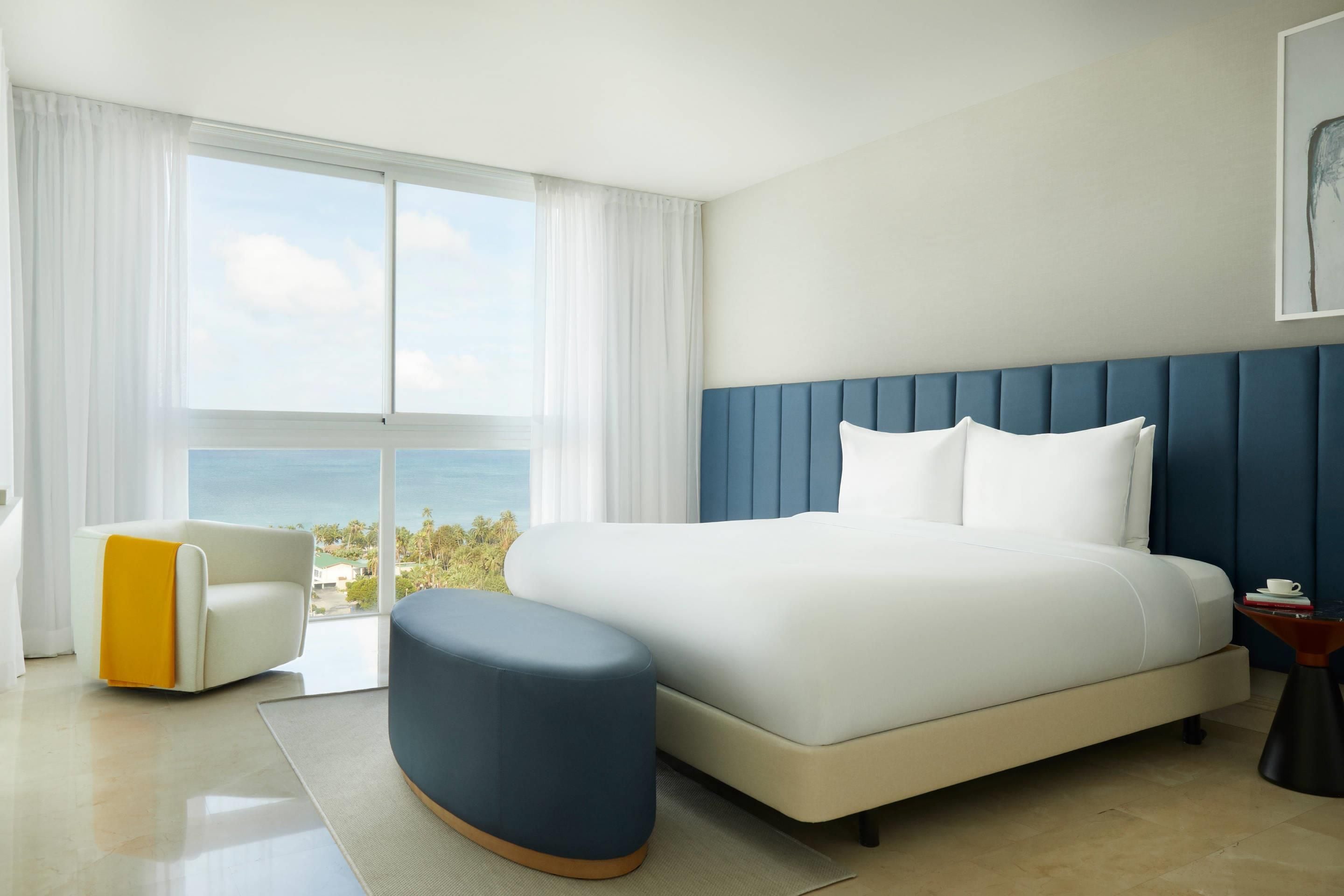 A modern hotel room with a large bed, a view of the ocean, a chair, and a small table with a cup.