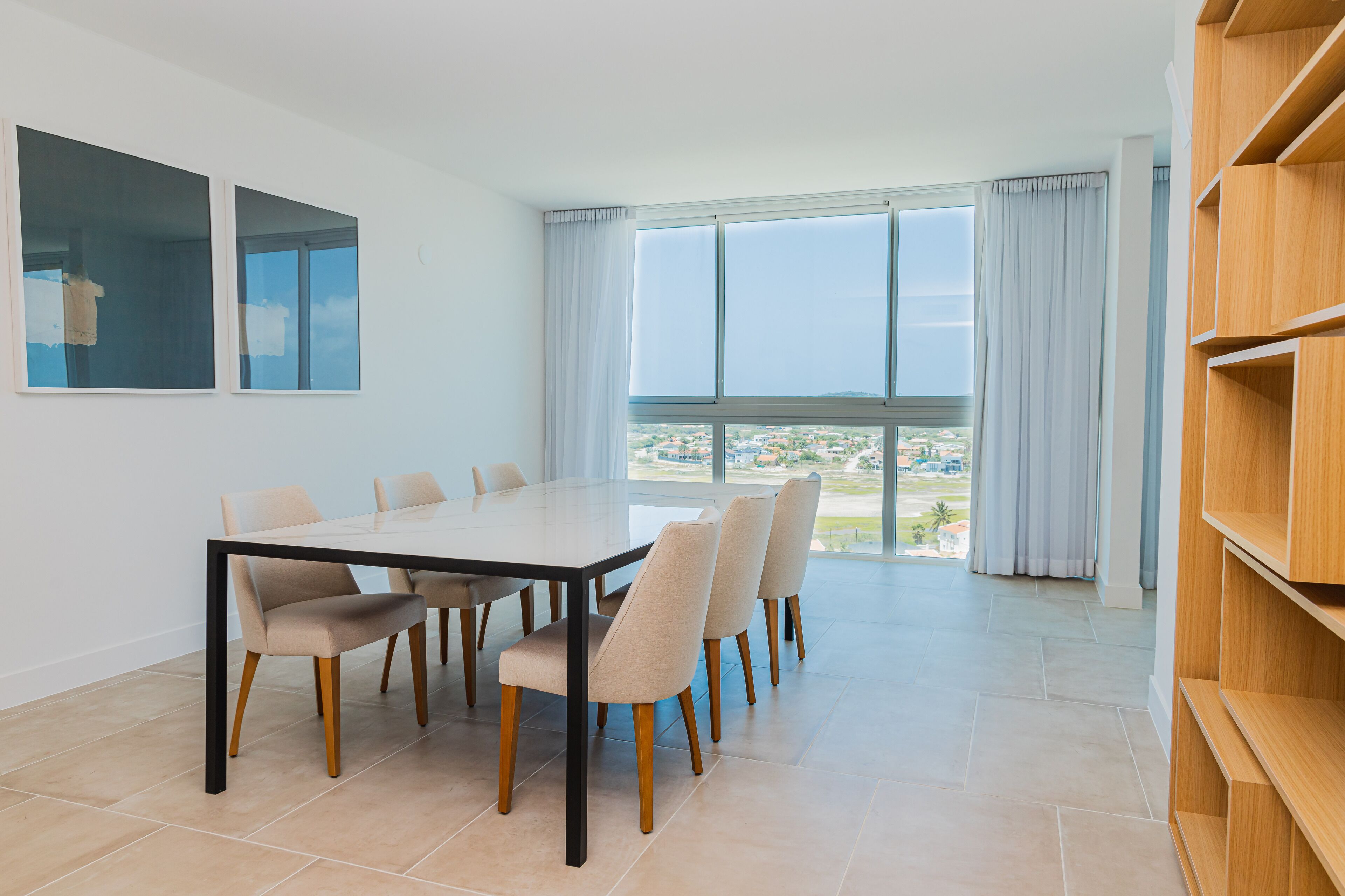 A modern dining area with a large table and six chairs, large windows with curtains, and a view of a cityscape.