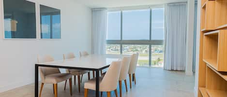 A modern dining area with a large table and six chairs, large windows with curtains, and a view of a cityscape.