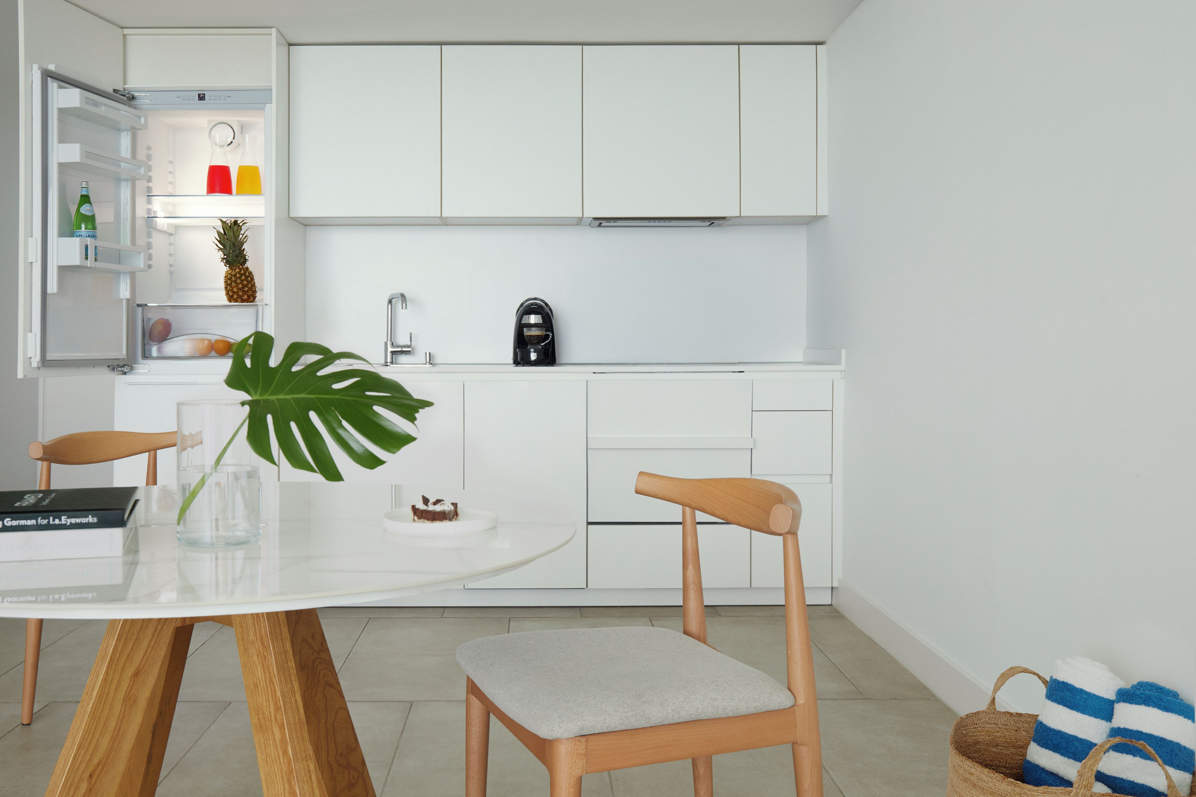 Une cuisine moderne avec des meubles blancs, une machine à café, une table à manger avec des chaises et une grande plante.