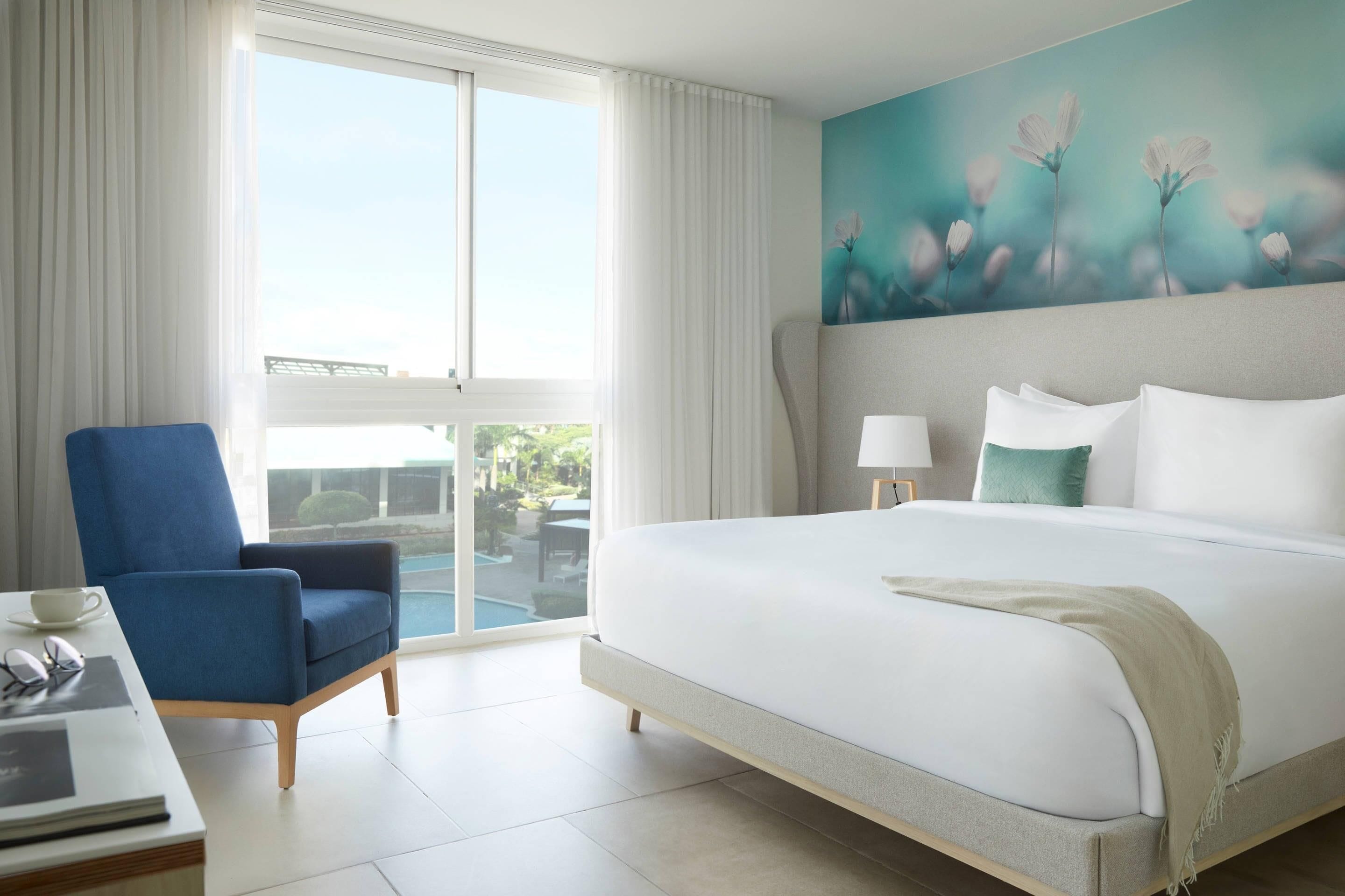 A hotel room with a large bed, a blue armchair, a desk with a laptop, and a painting of flowers on the wall.