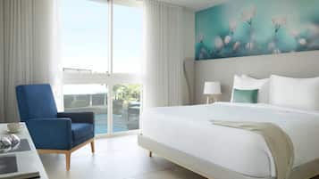 A hotel room with a large bed, a blue armchair, a desk with a laptop, and a painting of flowers on the wall.