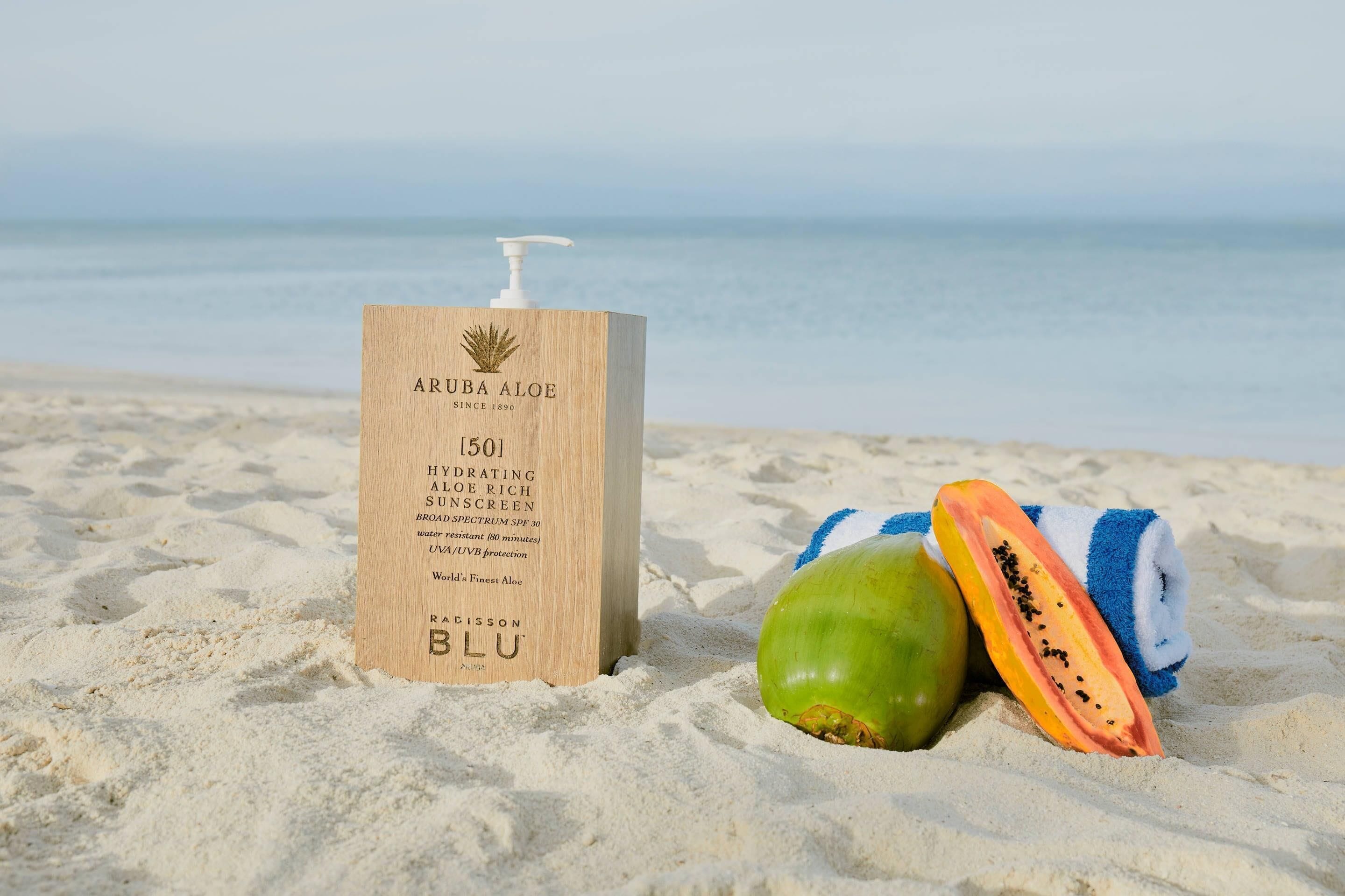 A bottle of Aruba Aloe hydrating aloe rich sunscreen on a sandy beach with a towel and a papaya.