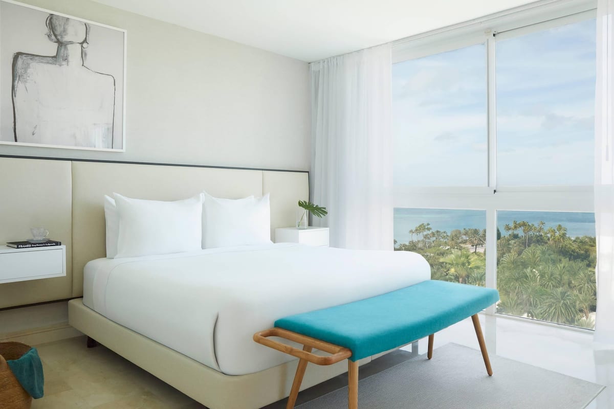 A modern hotel room with a large bed, a bench, a nightstand, a framed picture, and a view of the ocean.