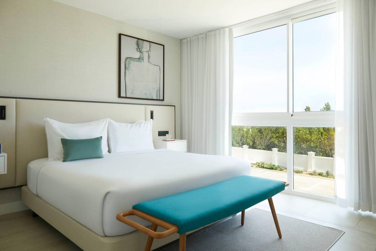 A modern hotel room with a large bed, a bench, a nightstand, a framed artwork, and a view of greenery through the window.