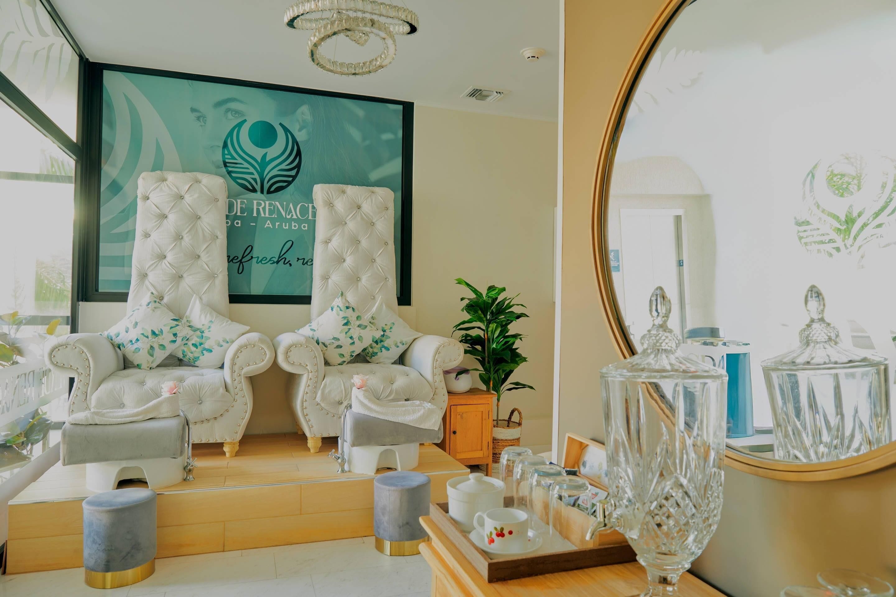 A spa room with two tufted chairs, a small table with a tray of items, a plant, and a large mirror.