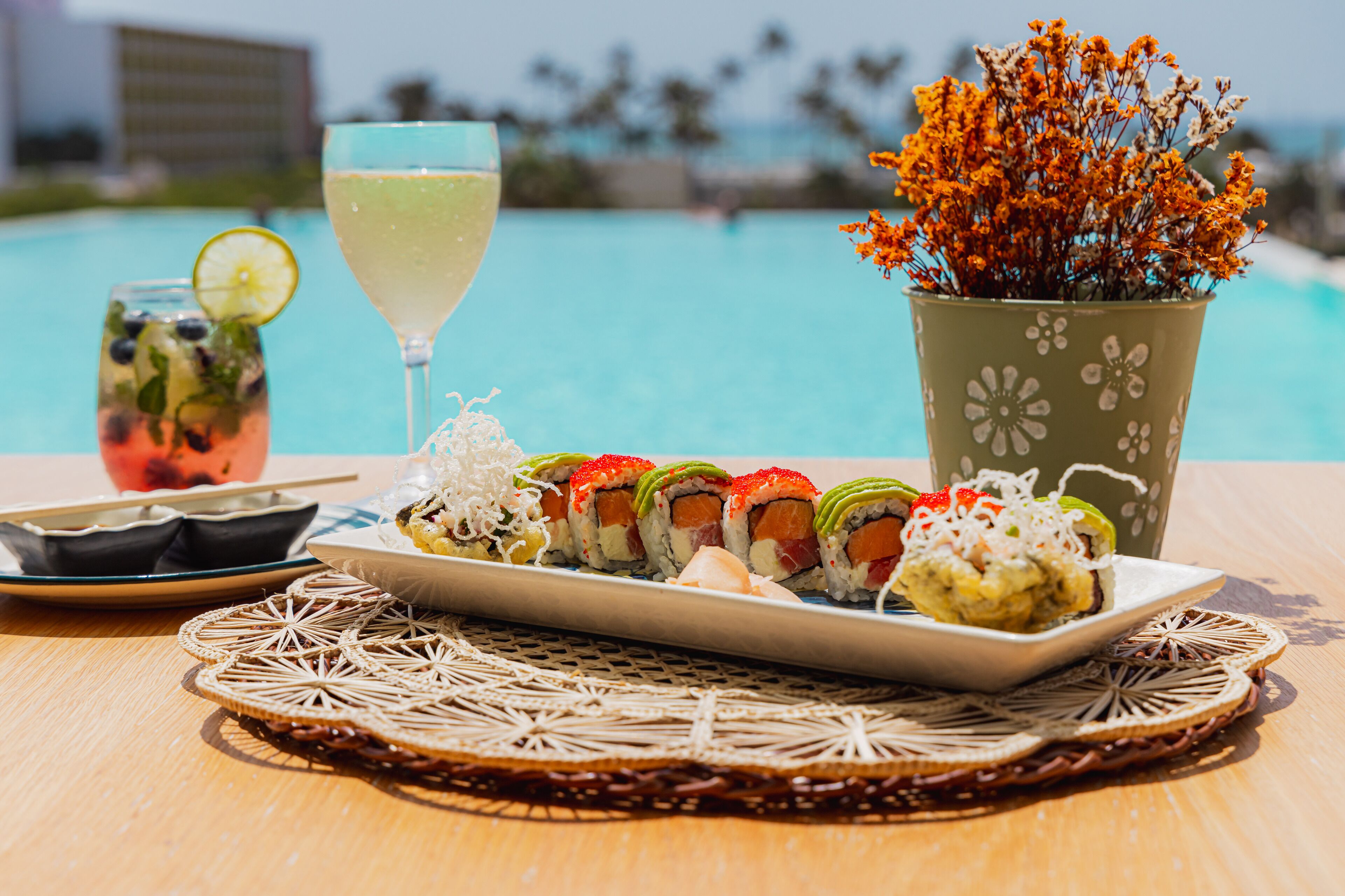 Una mesa con sushi, una bebida y un arreglo floral junto a una piscina.