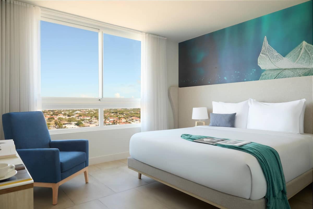 A hotel room with a large bed, a blue armchair, a view of a cityscape through a large window, and a painting of a sailboat on the wall.