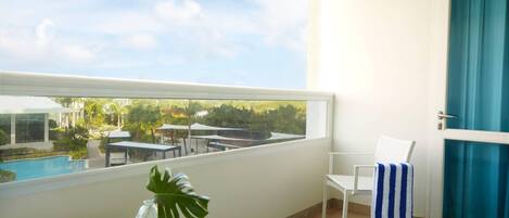 Un balcón con vistas a una piscina y follaje, una silla blanca, una toalla a rayas y una cesta de mimbre.