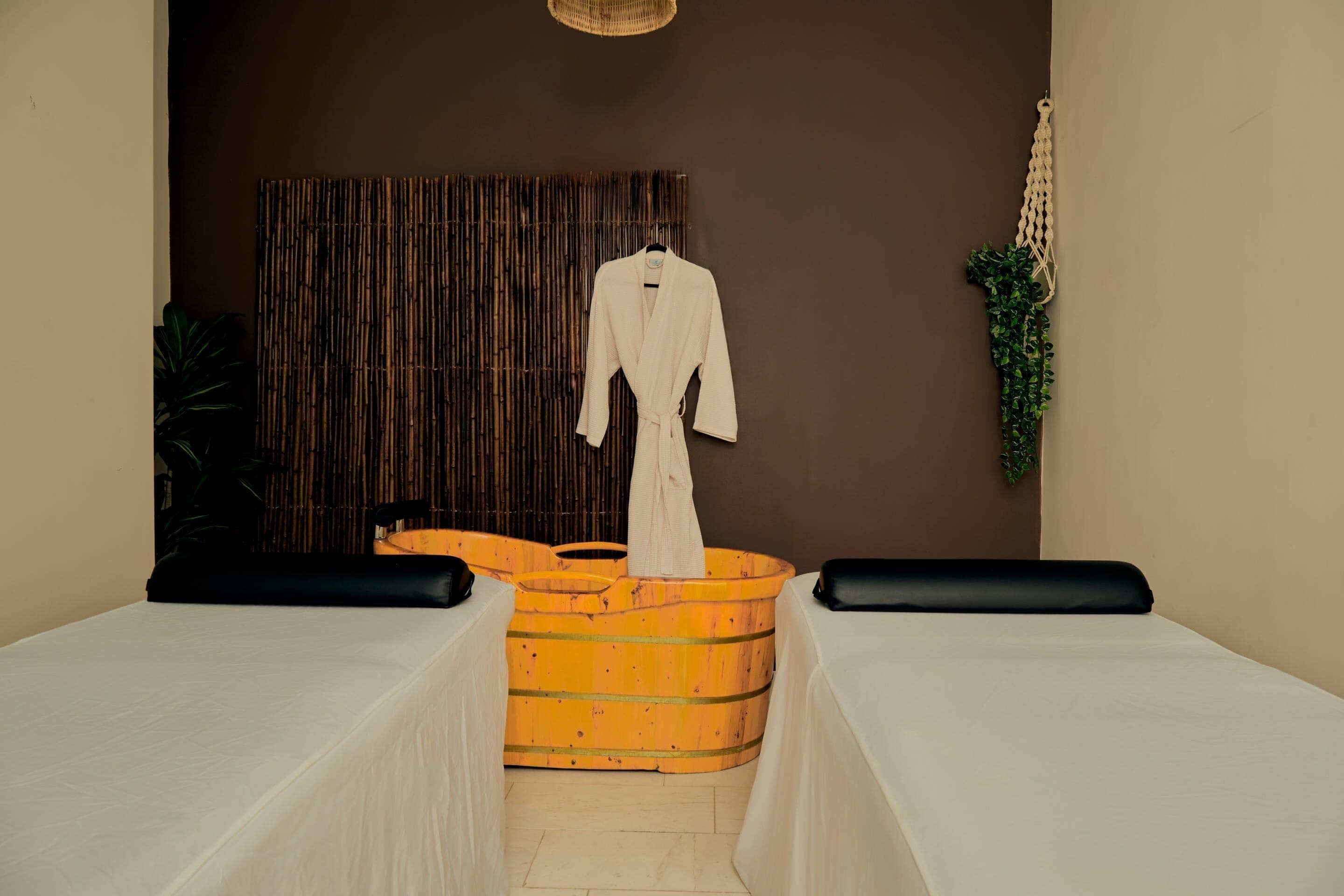 A spa room with a wooden tub, a white robe, and black cushions on the beds.