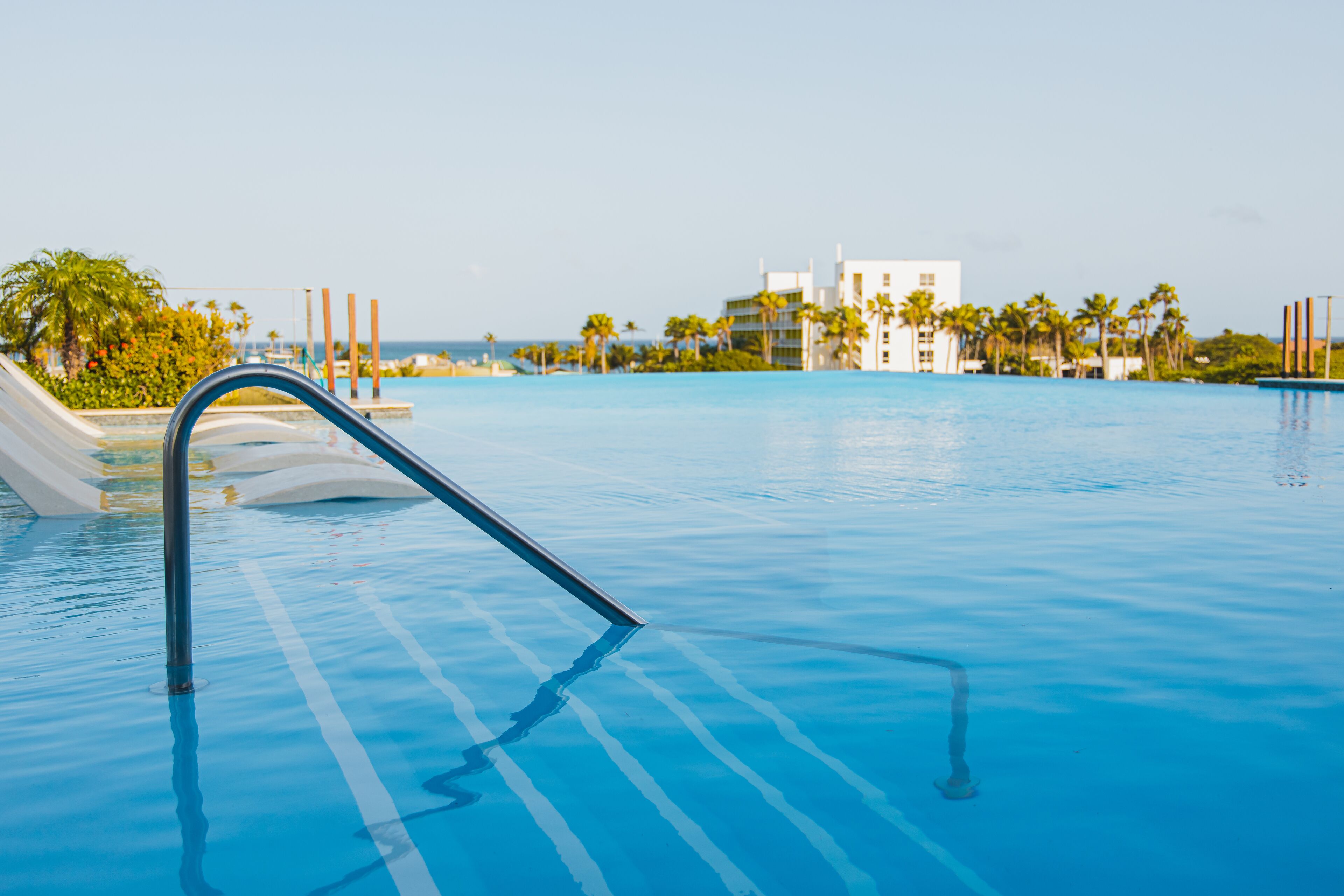 A swimming pool with clear blue water, a metal ladder, and a view of a resort building with palm trees in the background.