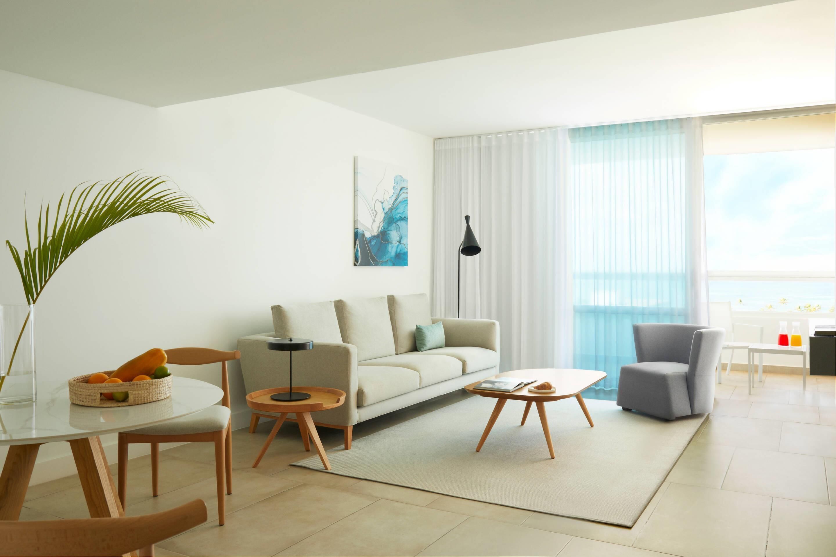 A modern living room with a sofa, coffee table, and chairs, a dining table with a vase of flowers, and a large window with curtains.
