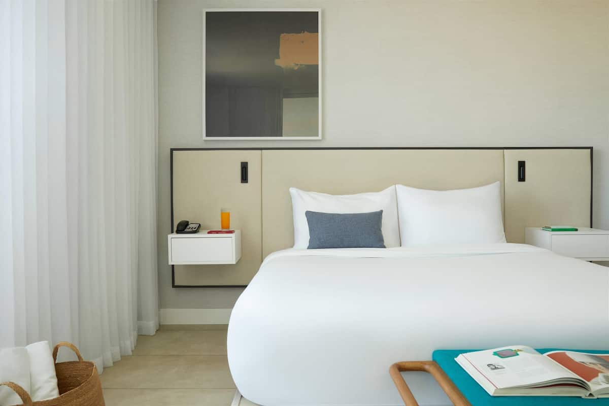 A modern hotel room with a large bed, a bedside table with a phone and a book, a small shelf with a bottle, and a framed picture on the wall.