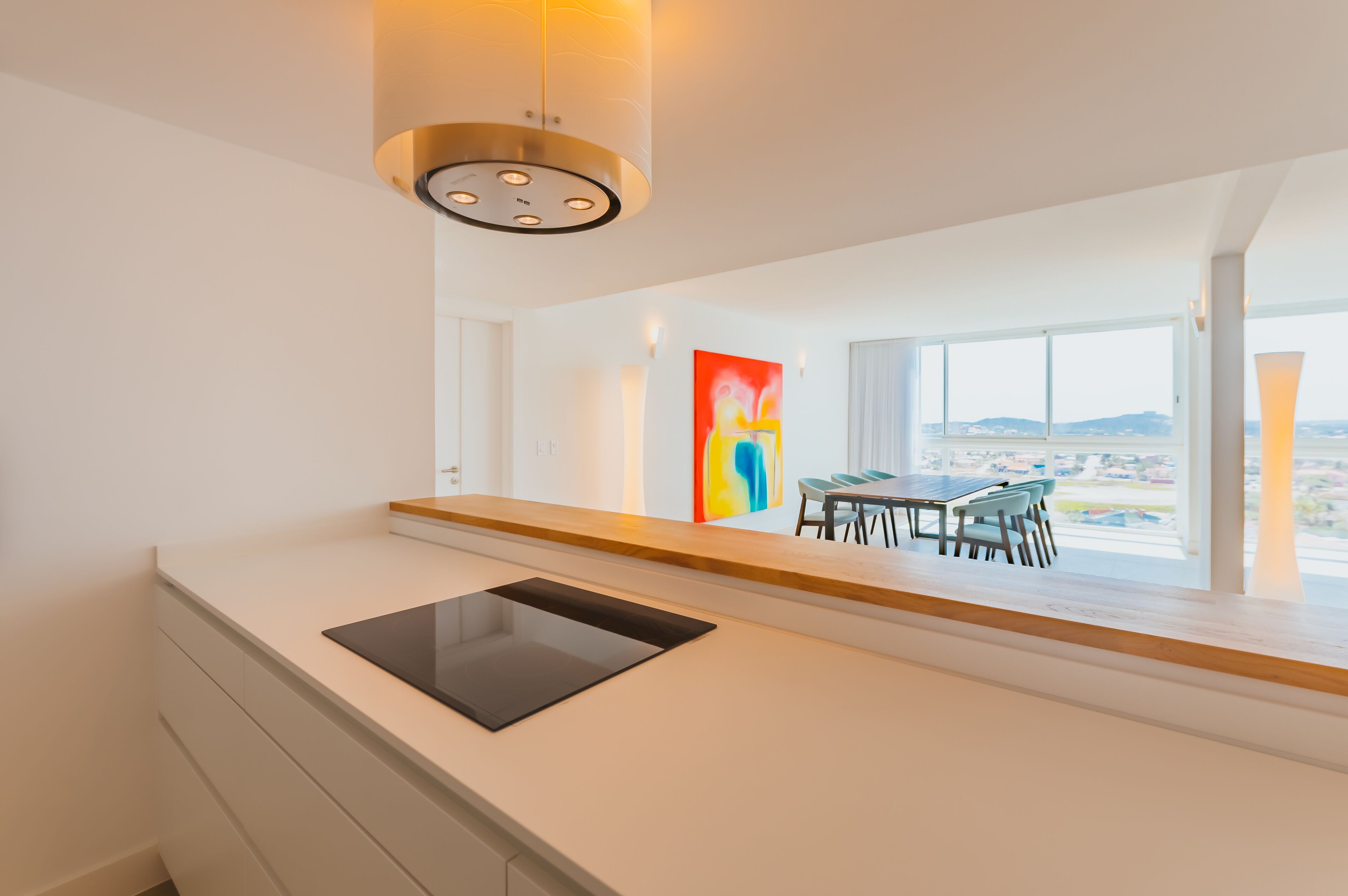 A modern kitchen with a built-in tablet, a dining area with a table and chairs, and a large window offering a view of the city.