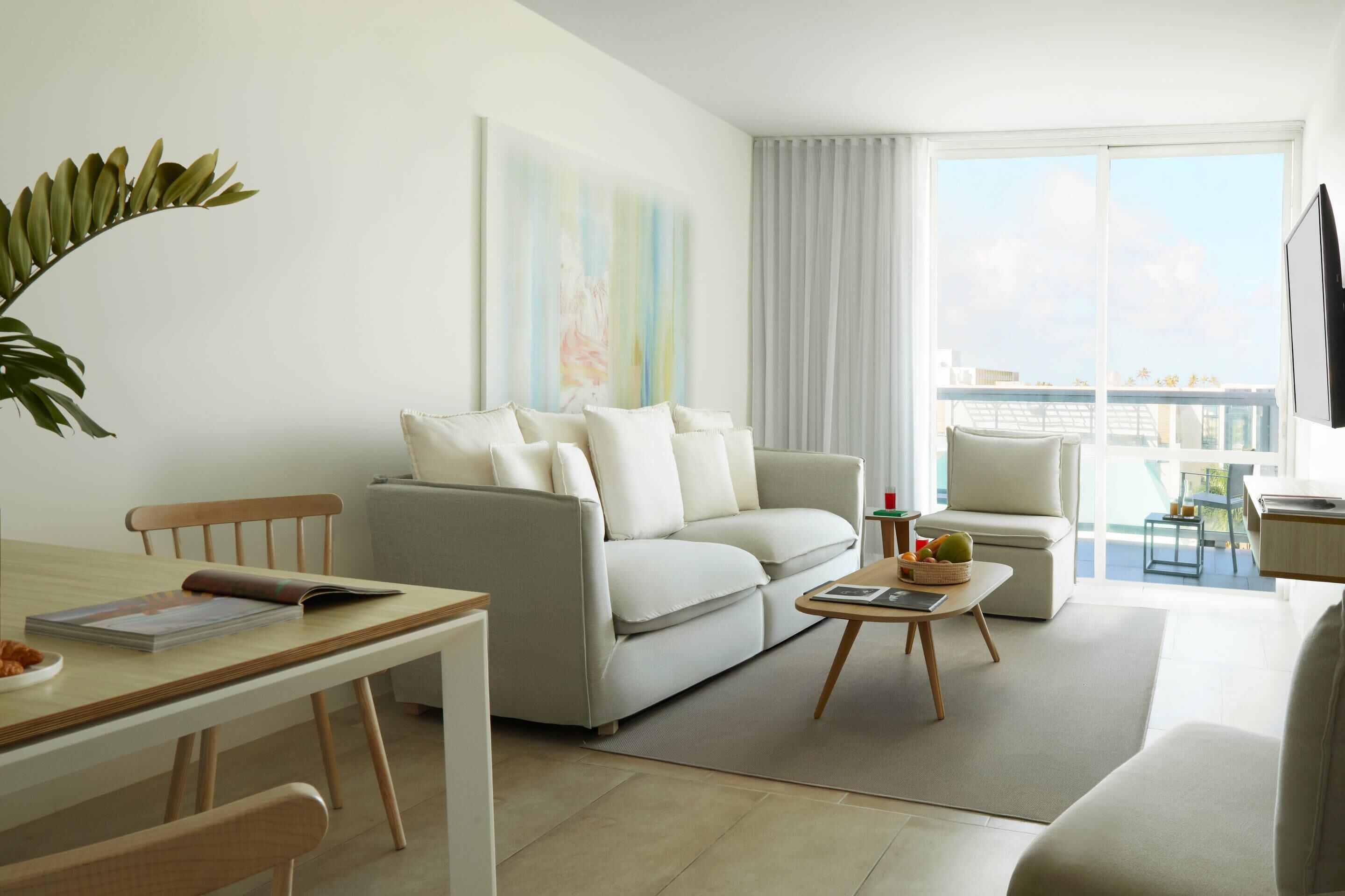 En moderne stue med sofa, spisebord og sofabord. Der er et stort vindue med gardiner, en plante og et billede på væggen.