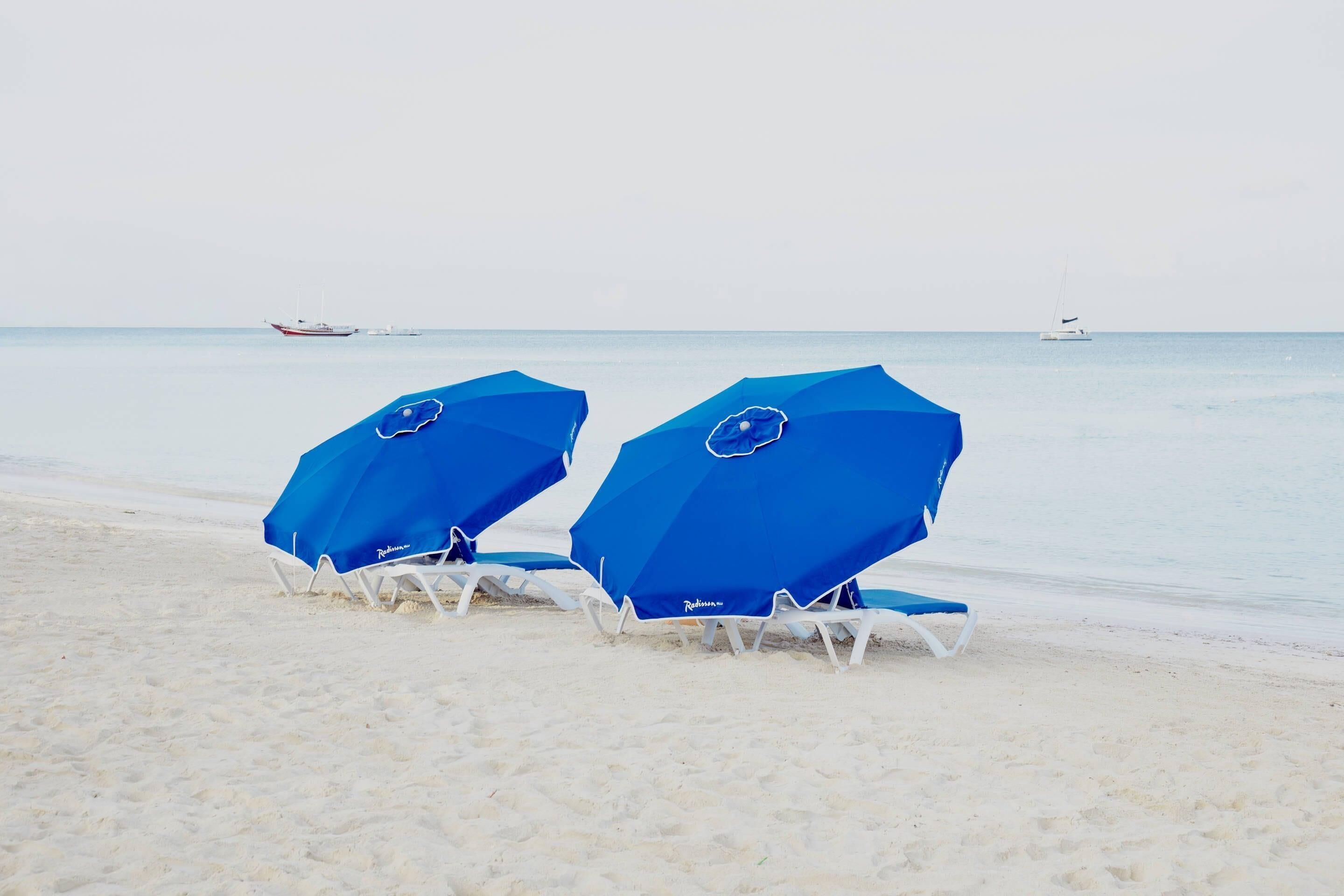 Deux parasols de plage bleus, accompagnés de chaises blanches, sont installés sur une plage de sable.