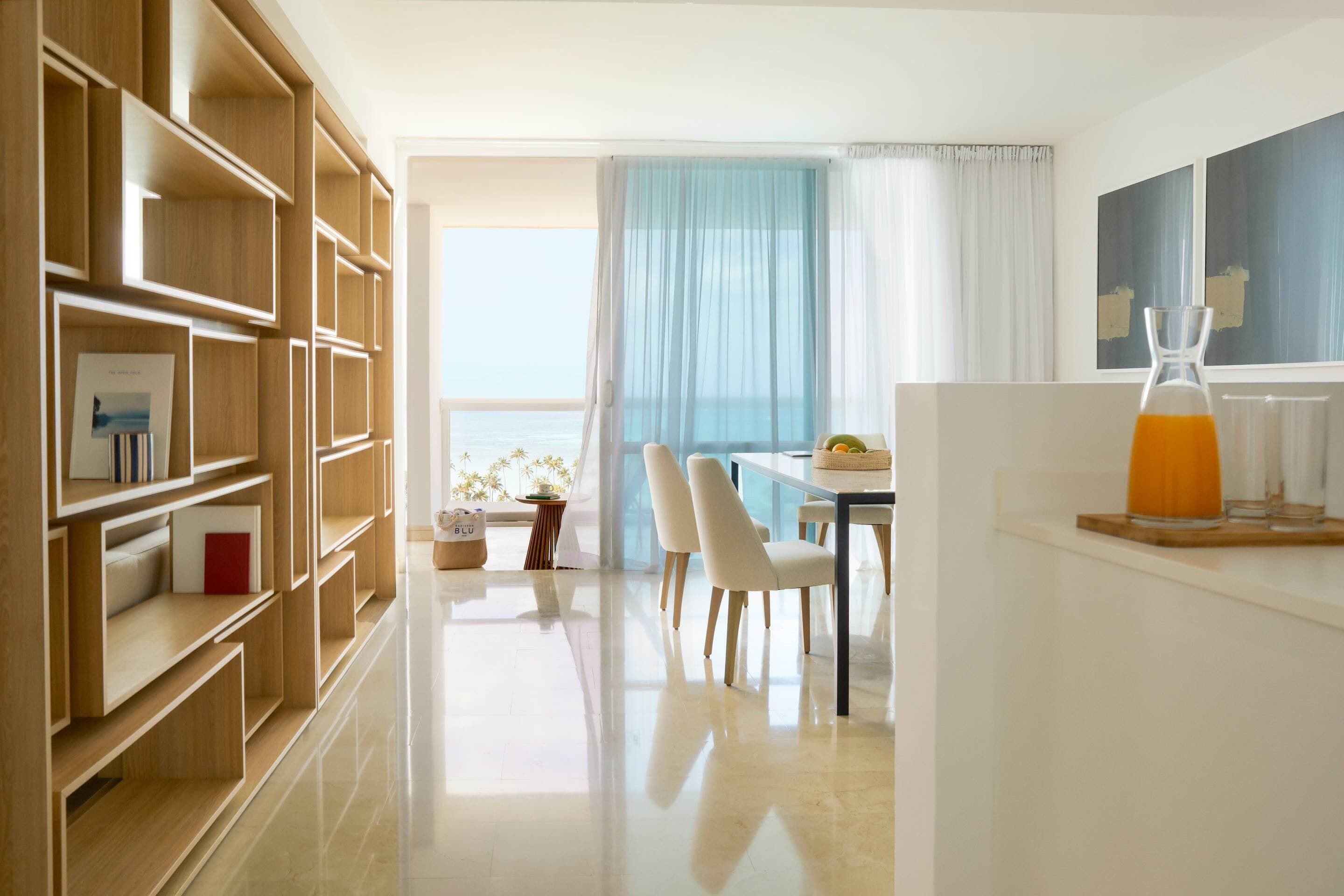 Ett modernt hotellrum med en bokhylla i trä, en matplats med bord och stolar samt utsikt över havet genom ett stort fönster.